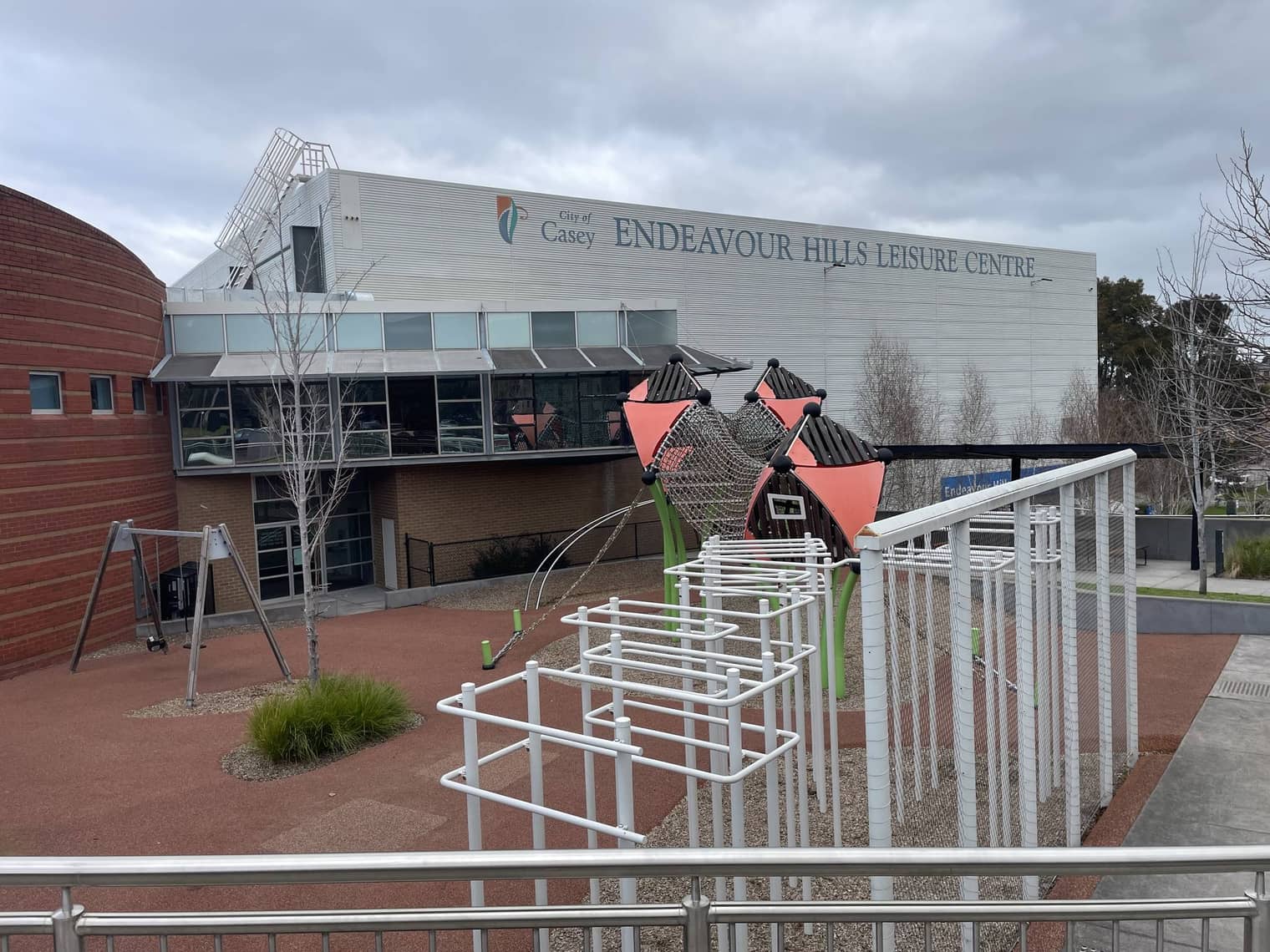 The Endeavour Hills Leisure Centre, a large pale-grey building, with a fenced children's playground and climbing net in the foreground.