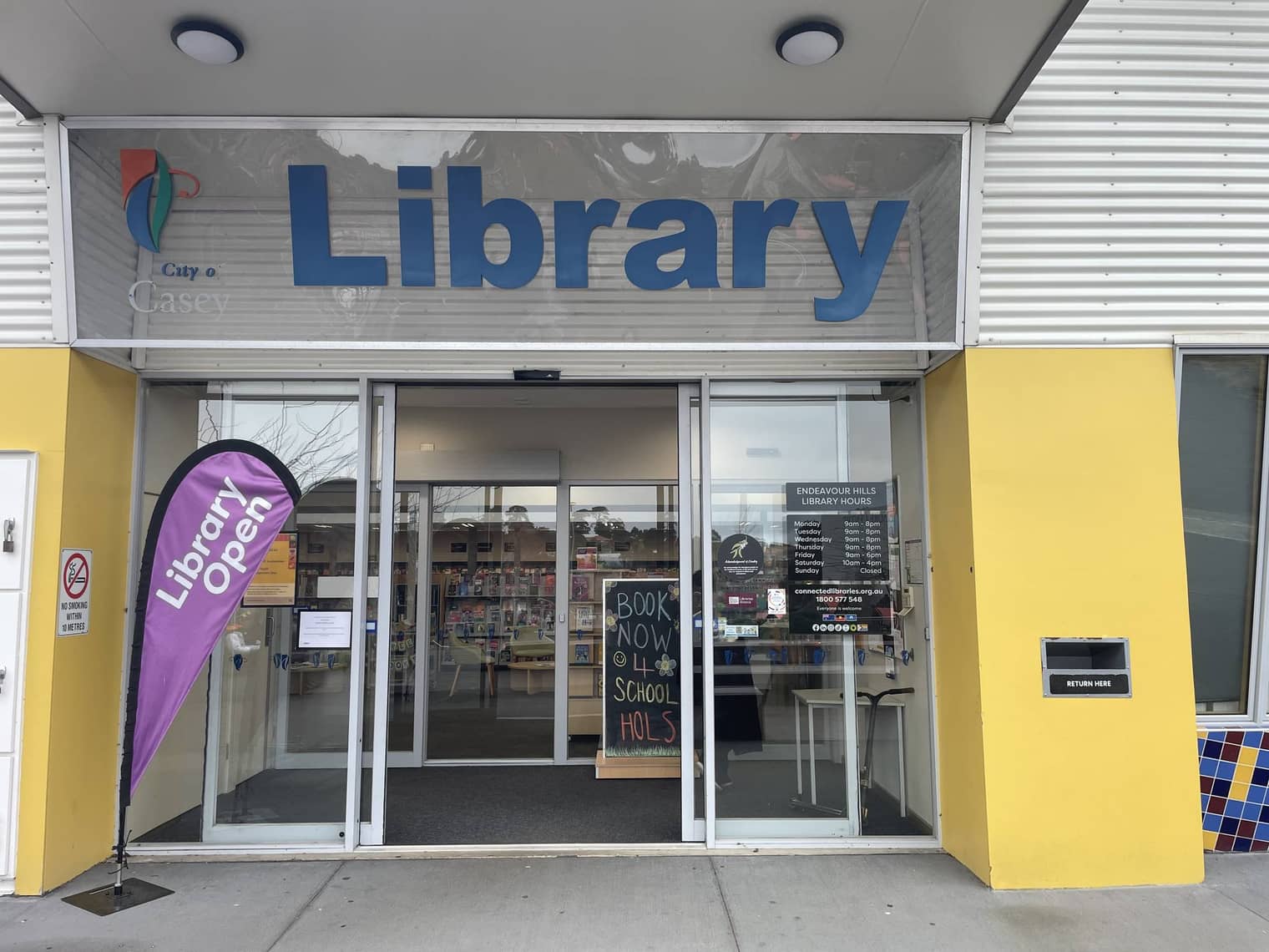 The glass front doors of Endeavour Hills Library, with a purple "Library Open" feather flag and a chalkboard advertising school-holiday programs.