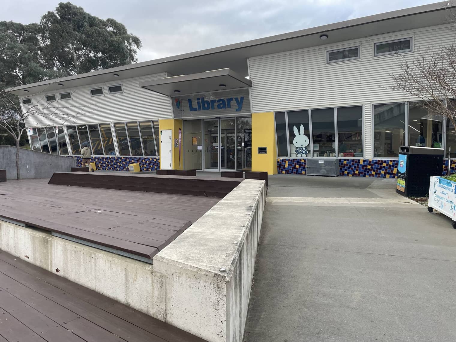 Exterior of Endeavour Hills Library, a long low white building with a raised timber deck out front, set in the civic precinct beside the shopping centre.