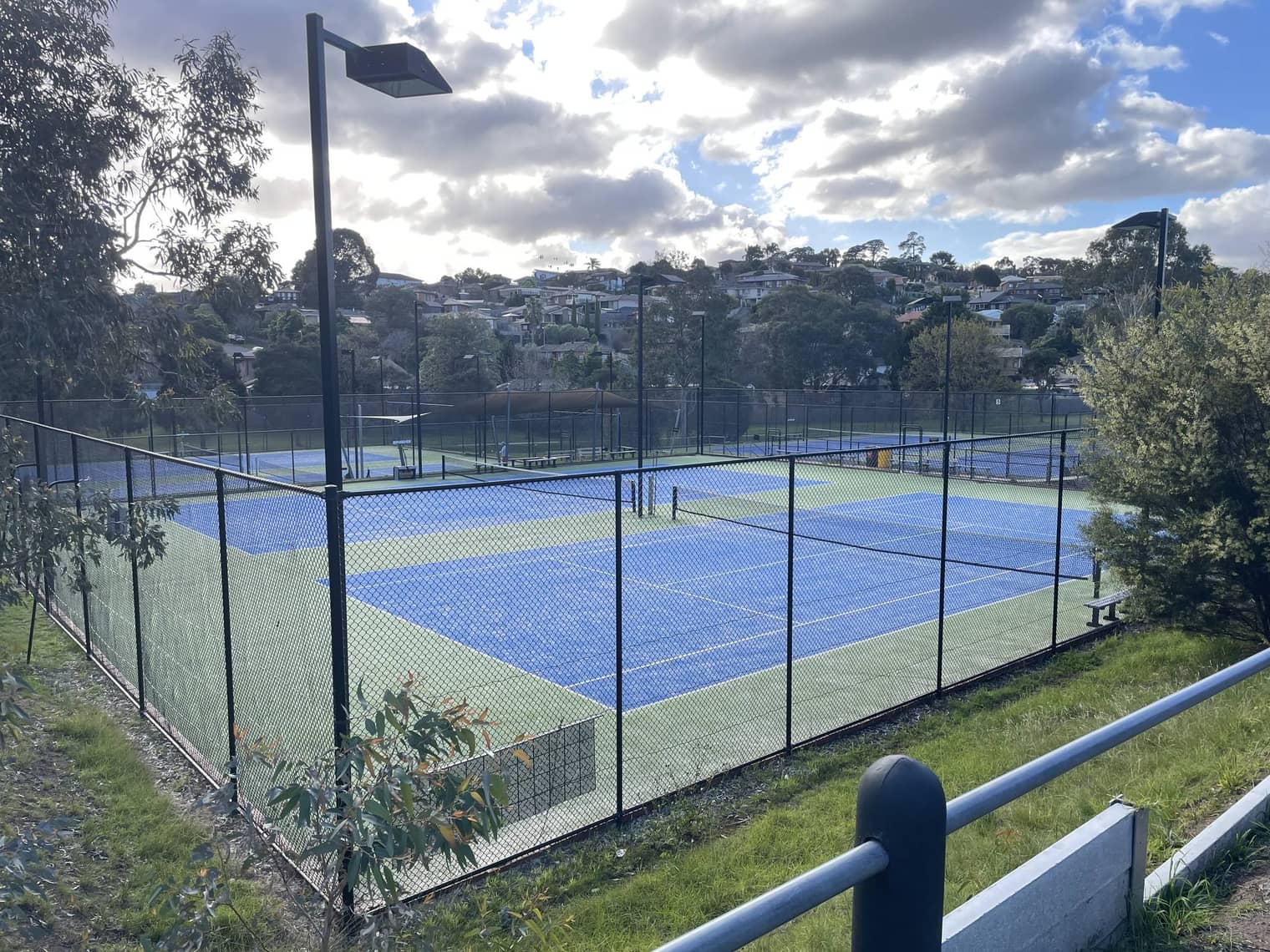 Blue-surfaced outdoor tennis courts at the Endeavour Hills Tennis Club — floodlit and fenced, with houses on the ridge behind.